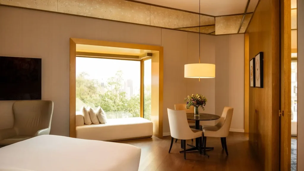 Modern Almanac Family Suite featuring a cozy bed, window seat with views of greenery, a round table with chairs, and a stylish hanging light fixture.